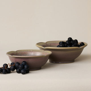 Two ceramic flower-shaped bowls with a muted mauve-pink and brown glaze, one filled with dark grapes and the other with black grapes, set against a warm beige background.