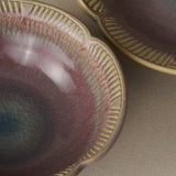Close-up of a ceramic bowl with a scalloped rim, featuring a textured glaze in dusty mauve, olive green, and blue-gray tones, with a second matching bowl partially visible in the background.