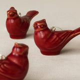 Three glossy red ceramic bird ornaments shaped like cardinals, each with a small metal hanging loop, arranged on a light beige fabric background.