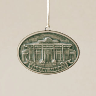 An oval Rookwood Findlay Market ceramic Christmas ornament in green Sencha glaze, featuring a relief design of Cincinnati's Findlay Market building with the text 'FINDLAY MARKET' on a banner, hanging from a white ribbon against a textured beige background.