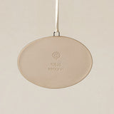 Backside of the Rookwood Findlay Market ornament in Sencha glaze: oval light beige ceramic with an embossed RP logo, item number, and 'MMXXVI' in the center, suspended by a white ribbon against a textured beige background.