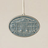 An oval Rookwood Findlay Market ceramic Christmas ornament in blue Teton glaze, featuring a relief design of Cincinnati's Findlay Market building with the text 'FINDLAY MARKET' on a banner, hanging from a white ribbon against a textured beige background.