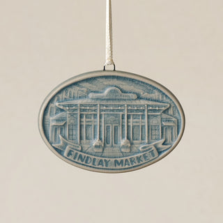 An oval Rookwood Findlay Market ceramic Christmas ornament in blue Teton glaze, featuring a relief design of Cincinnati's Findlay Market building with the text 'FINDLAY MARKET' on a banner, hanging from a white ribbon against a textured beige background.