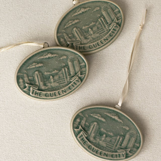Three oval Rookwood Queen City ceramic Christmas ornaments in green Sencha glaze, featuring a relief design of the Cincinnati, Ohio skyline with the text 'THE QUEEN CITY' on a banner, lying on a textured beige surface with white ribbons.