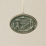 An oval Rookwood Queen City ceramic Christmas ornament in green Sencha glaze, featuring a relief design of the Cincinnati, Ohio skyline with the text 'THE QUEEN CITY' on a banner, hanging from a white ribbon against a textured beige background.