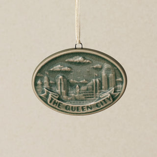An oval Rookwood Queen City ceramic Christmas ornament in green Sencha glaze, featuring a relief design of the Cincinnati, Ohio skyline with the text 'THE QUEEN CITY' on a banner, hanging from a white ribbon against a textured beige background.
