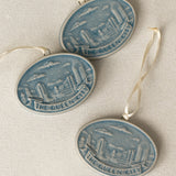 Three oval ceramic Christmas ornament featuring a blue and white relief design of the Cincinnati, Ohio skyline with the text 'THE QUEEN CITY' on a banner, hanging from a white ribbon against a textured beige background.