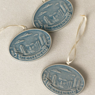 Three oval ceramic Christmas ornament featuring a blue and white relief design of the Cincinnati, Ohio skyline with the text 'THE QUEEN CITY' on a banner, hanging from a white ribbon against a textured beige background.
