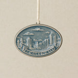 An oval ceramic Christmas ornament featuring a blue and white relief design of the Cincinnati, Ohio skyline with the text 'THE QUEEN CITY' on a banner, hanging from a white ribbon against a textured beige background.