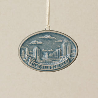 An oval ceramic Christmas ornament featuring a blue and white relief design of the Cincinnati, Ohio skyline with the text 'THE QUEEN CITY' on a banner, hanging from a white ribbon against a textured beige background.