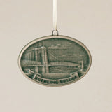 An oval Rookwood Roebling Bridge ceramic Christmas ornament in green Sencha glaze, featuring a relief design of Cincinnati's Roebling Suspension Bridge with the text 'ROEBLING BRIDGE' on a banner, hanging from a white ribbon against a textured beige background.