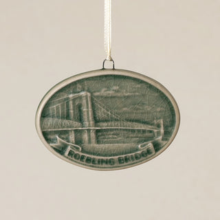An oval Rookwood Roebling Bridge ceramic Christmas ornament in green Sencha glaze, featuring a relief design of Cincinnati's Roebling Suspension Bridge with the text 'ROEBLING BRIDGE' on a banner, hanging from a white ribbon against a textured beige background.