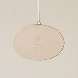 Backside of the Rookwood Roebling Bridge ornament in Sencha glaze: oval light beige ceramic with an embossed RP logo, item number, and 'MMXXVI' in the center, suspended by a white ribbon against a textured beige background.