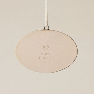 Backside of the Rookwood Roebling Bridge ornament in Sencha glaze: oval light beige ceramic with an embossed RP logo, item number, and 'MMXXVI' in the center, suspended by a white ribbon against a textured beige background.