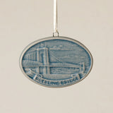 An oval Rookwood Roebling Bridge ceramic Christmas ornament in blue Teton glaze, featuring a relief design of Cincinnati's Roebling Suspension Bridge with the text 'ROEBLING BRIDGE' on a banner, hanging from a white ribbon against a textured beige background.