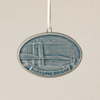An oval Rookwood Roebling Bridge ceramic Christmas ornament in blue Teton glaze, featuring a relief design of Cincinnati's Roebling Suspension Bridge with the text 'ROEBLING BRIDGE' on a banner, hanging from a white ribbon against a textured beige background.