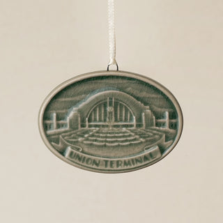 An oval Rookwood Union Terminal ceramic Christmas ornament in green Sencha glaze, featuring a relief design of Cincinnati's Union Terminal with the text 'UNION TERMINAL' on a banner, hanging from a white ribbon against a textured beige background.