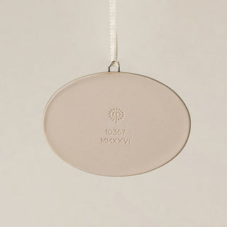 Backside of the Rookwood Union Terminal ornament in Sencha glaze: oval light beige ceramic with an embossed RP logo, item number, and 'MMXXVI' in the center, suspended by a white ribbon against a textured beige background.