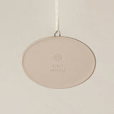Backside of the Rookwood Union Terminal ornament in Teton glaze: oval light beige ceramic with an embossed RP logo, item number, and 'MMXXVI' in the center, suspended by a white ribbon against a textured beige background.