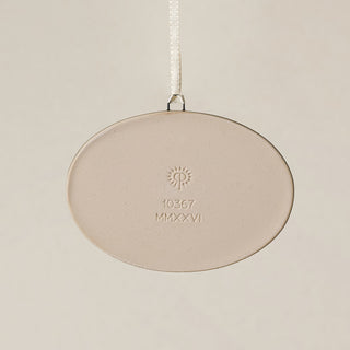 Backside of the Rookwood Union Terminal ornament in Teton glaze: oval light beige ceramic with an embossed RP logo, item number, and 'MMXXVI' in the center, suspended by a white ribbon against a textured beige background.