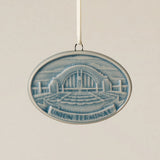 An oval Rookwood Union Terminal ceramic Christmas ornament in blue Teton glaze, featuring a relief design of Cincinnati's Union Terminal with the text 'UNION TERMINAL' on a banner, hanging from a white ribbon against a textured beige background.