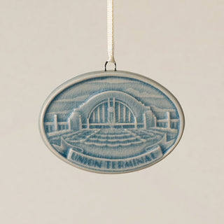 An oval Rookwood Union Terminal ceramic Christmas ornament in blue Teton glaze, featuring a relief design of Cincinnati's Union Terminal with the text 'UNION TERMINAL' on a banner, hanging from a white ribbon against a textured beige background.