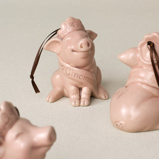 A cluster of pink ceramic pig ornaments, wearing a crown and a sash that reads 'Cincinnati,' paired with a brown ribbon against a plain light beige background.