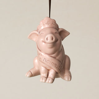 A pink ceramic pig ornament, wearing a crown and a sash that reads 'Cincinnati,' hanging from a brown ribbon against a plain light beige background.